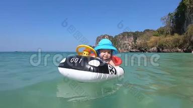 在开阔的大海和群山的背景下，一个穿着蓝色巴拿马的快乐宝宝在一艘充气汽车形状的床垫船上游泳。